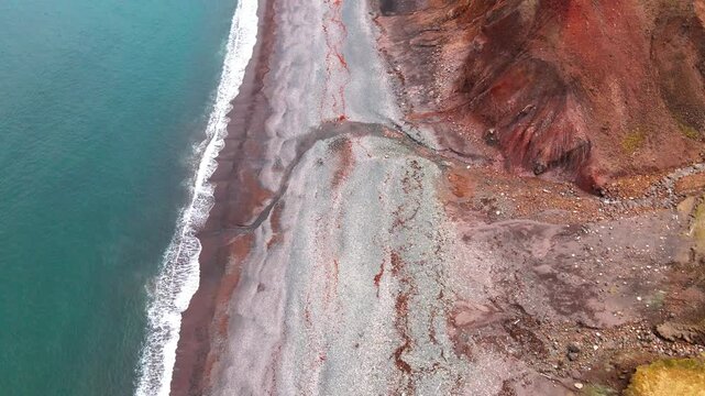 Aerial view of the colorful coastline of Brunavik with reddish cliffs meeting the turquoise ocean waves on a pebble beach in Bakkagerdi, Mulathing, Iceland.