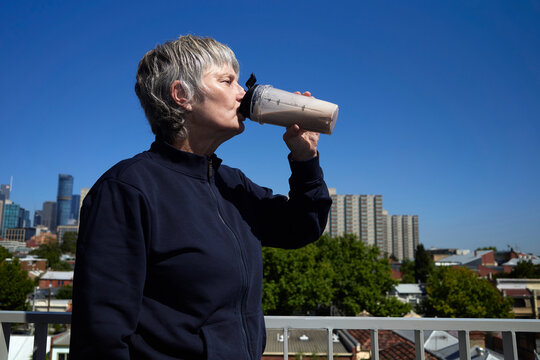 Woman Drinks a Protein Shake While Standing on a Balcony in the City