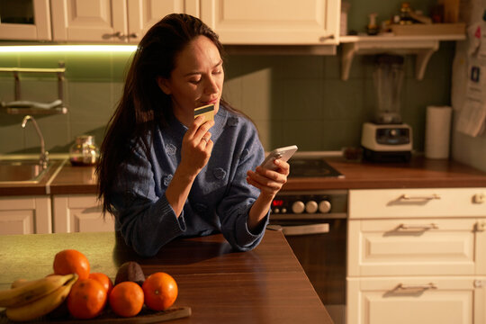 Woman making online payment by card at sunset in kitchen