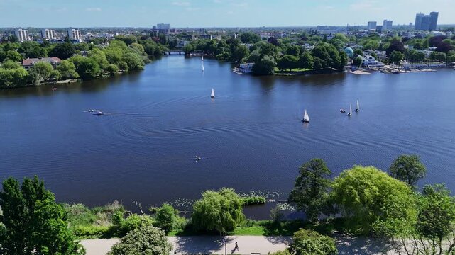 Aerial view of the Aussenalster lake with sailboats and rowers surrounded by lush green trees and urban skyline under a clear blue sky in Hamburg, Hamburg, Germany.
