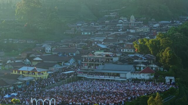 Aerial view of a large crowd gathered for Eid Al-Adha prayer in an open field surrounded by village houses and misty hills in Kecamatan Kalikajar, Jawa Tengah, Indonesia.
