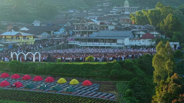 Aerial view of a large crowd gathered for Eid Al-Adha Prayer in Garung with surrounding buildings and hills in Kecamatan Kalikajar, Jawa Tengah, Indonesia.