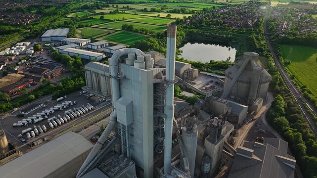 Aerial view of the Rugby Cement Plant featuring tall chimneys, industrial silos, and parked trucks surrounded by green fields and a pond in Rugby, England, United Kingdom.