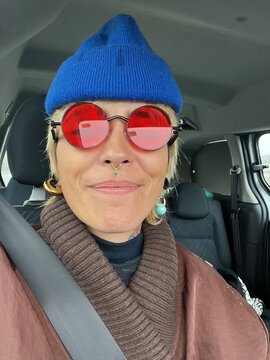 Person sits in car wearing sunglasses and a hat while smiling.