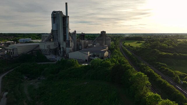 Aerial view of Rugby Cement Plant with its tall chimney and industrial structures next to a railway line and green fields during sunset Rugby, England, United Kingdom.