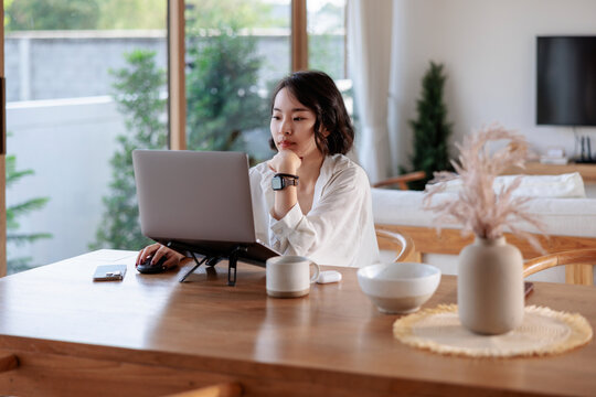 Woman working on laptop at home, thoughtfully looking at screen.
