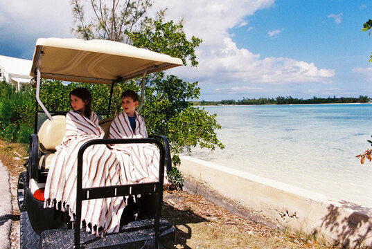 Siblings on vacation sit in golf cart wrapped up in beach towels 