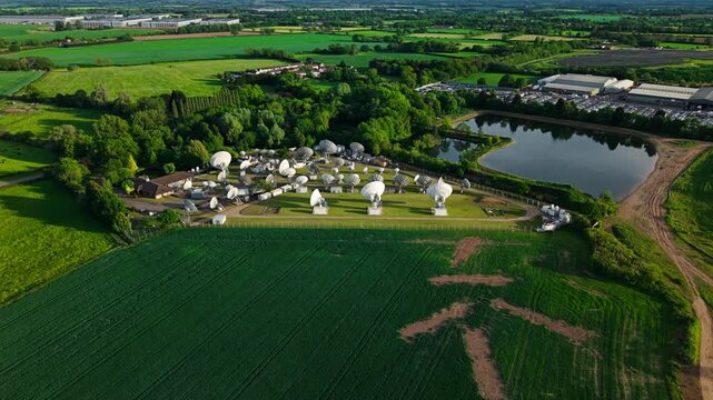 Aerial view of Satellite Mediaport Services with multiple satellite dishes surrounded by green fields and a small pond in Long Lawford, England, United Kingdom.