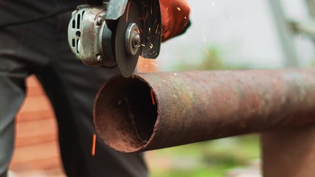 Worker in protective gloves uses electric grinder on rusty steel pipe outdoors. Bright sparks and powerful cutting tool create dynamic industrial scene focused on repair and metal preparation.