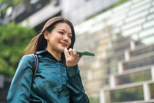 Smiling asian business woman using AI voice assistant on smartphone outdoors, Smart agent technology, Female corporate employee recording audio message or speaking to artificial intelligence app
