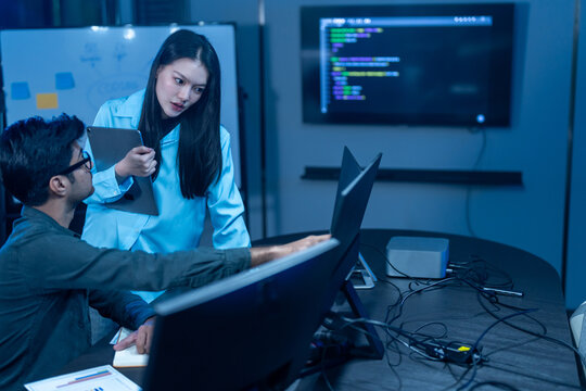 Asian female manager talking to male programmer writing code in dark blue office, Tech team discussing software development, cybersecurity and AI technology, Late night working business team concepts
