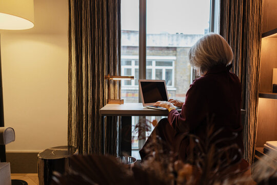 Senior woman working on laptop near window in hotel room