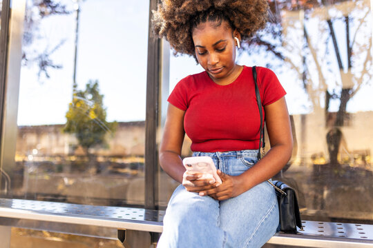 Black woman using her phone while waiting at a bus stop.