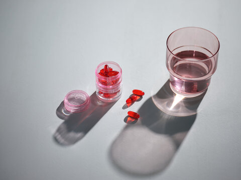 Red capsules and a glass of water on a light gray surface with shadows