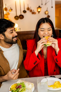 Woman eating hamburger at restaurant with friend. Direct flash
