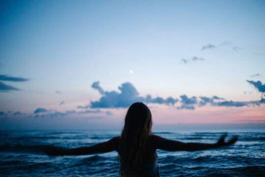 Woman facing ocean with outstretched arms at blue hour