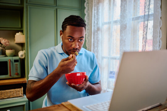Young Black Man Eating Food while Working on Laptop in Kitchen