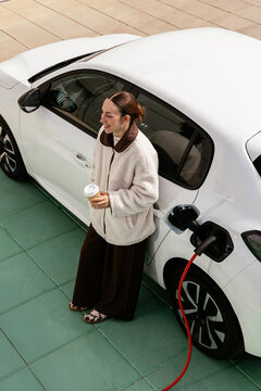 Woman charging electric car and holding coffee cup