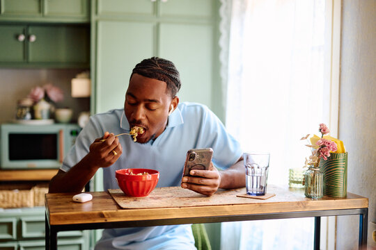 Young Black Man Eating Food while Checking Smartphone at Kitchen Table