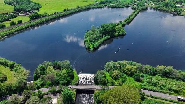 Aerial view of Kingsbury Water Park featuring a small green island, a weir with flowing water, and a road bridge surrounded by lush trees Kingsbury, England, United Kingdom.
