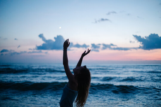 Woman reaching toward moon above ocean at blue hour