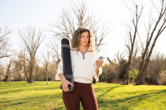 Woman checks her phone while holding yoga mat in a spring park