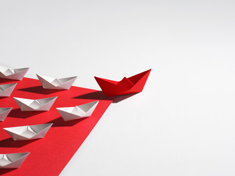 Red origami boat leads a fleet of white paper boats on a red path
