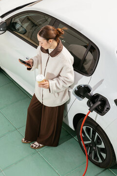 Woman charging electric car while using smartphone and drink coffee