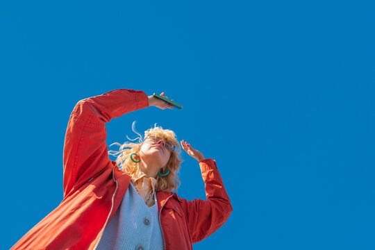 Low angle view of dancing woman holding phone under blue sky