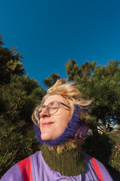 Woman in colorful knit hood enjoying sunlight by pine trees