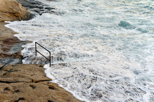 Huge wave crashes over rocky plateau, railing barely visible
