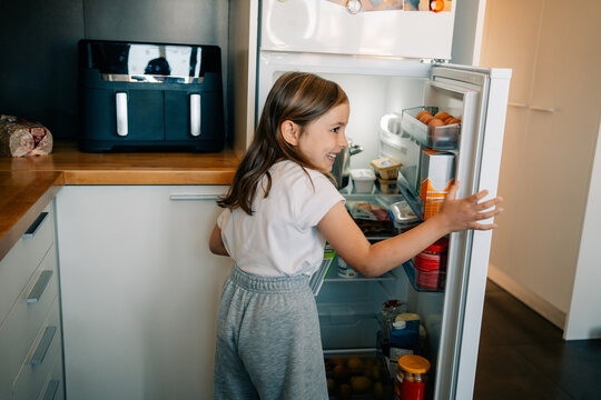Girl Looks Inside Refrigerator in Modern Kitchen While Smiling