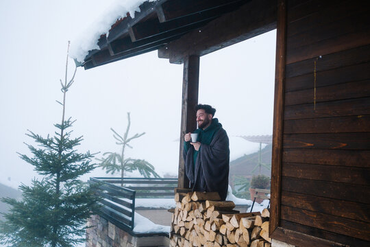 Man wrapped in blanket with hot drink on snowy mountain cabin terrace