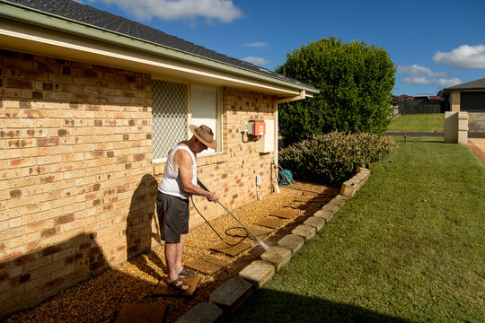 Elderly man pressure washing garden edge beside house.
