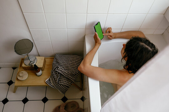 A person is taking a bath and scrolling a phone with a green screen