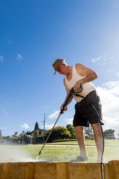 Elderly man pressure washing concrete blocks under blue sky
