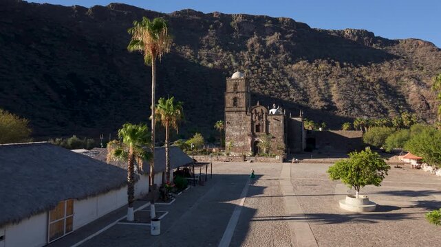 Watch aerial views of San Javier Mission at sunrise showing the town, palm trees, and mountains in Baja California Sur, Mexico