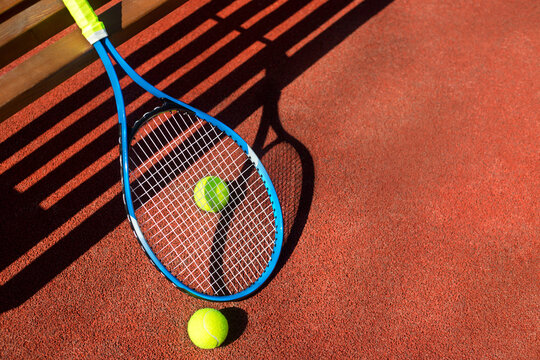 Tennis racket and two balls near a bench on a tennis court.
