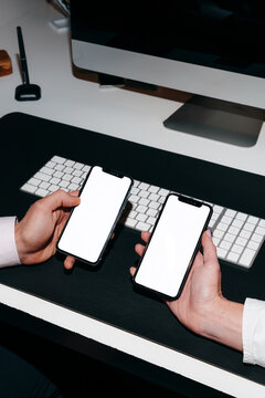 Two Hands Holding Smartphones With White Screen on the Desk