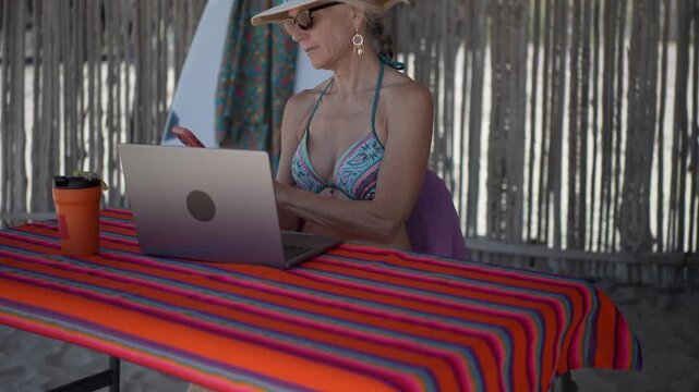 Mature woman using a computer and phone under a palapa on a beach during vacation time in a tropical setting.