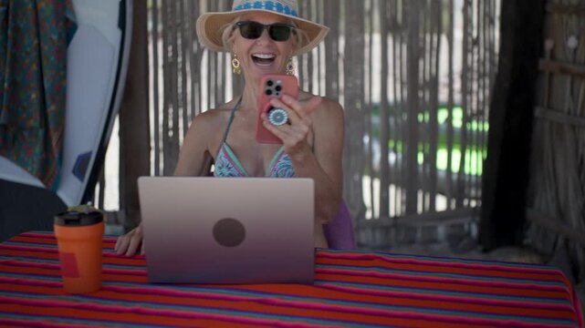 Happy mature woman works with computer and has video chat on her phone under a palapa on a tropical beach while enjoying her vacation under the sun.