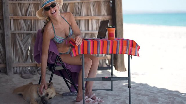 Happy mature woman sits at table on beach with computer and dog while enjoying drinks and working on vacation in tropical setting.