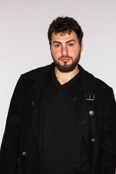 Man in black coat stands against a plain background indoors