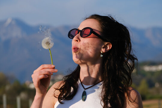 Dark-haired girl blowing a dandelion in a white t-shirt.