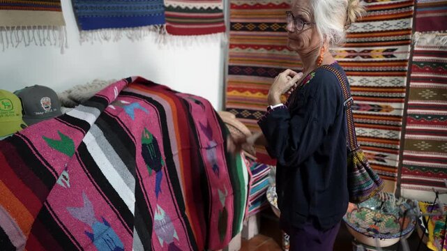 Senior woman explores a tourist shop in Mulege looking at colorful blankets and handmade items with interest and curiosity.