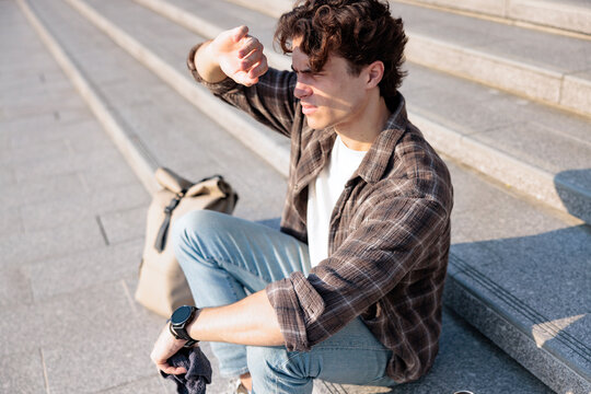 Man on steps shields eyes from scorching sun, backpack nearby.