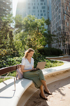 A woman uses a laptop while sitting on a bench in a city park