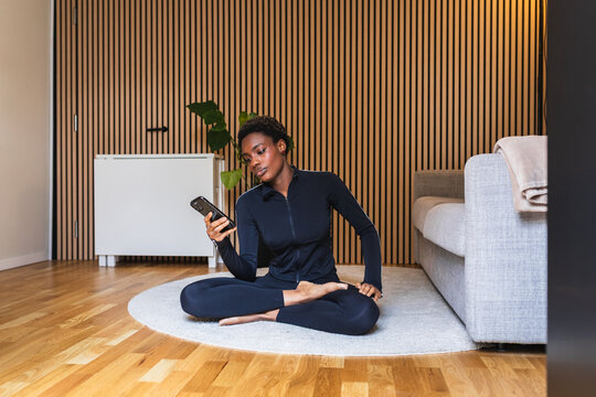 African Woman Checking Phone During Yoga in Small Apartment
