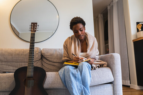 Young Woman Writing in Notebook with Guitar on Sofa