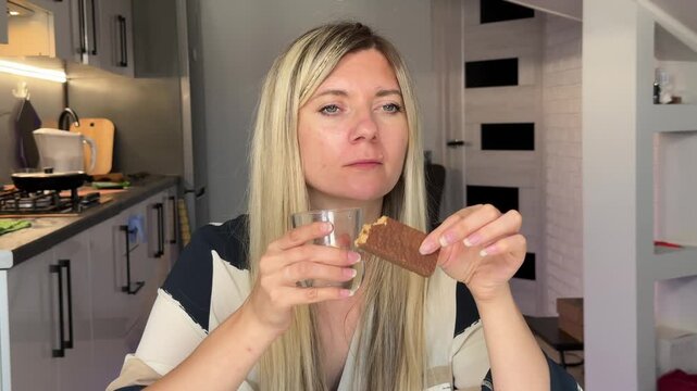Fairskinned Woman Innocently Tastes Chocolate Bar While Casually Reviewing Her Snack In Cozy Kitchen
